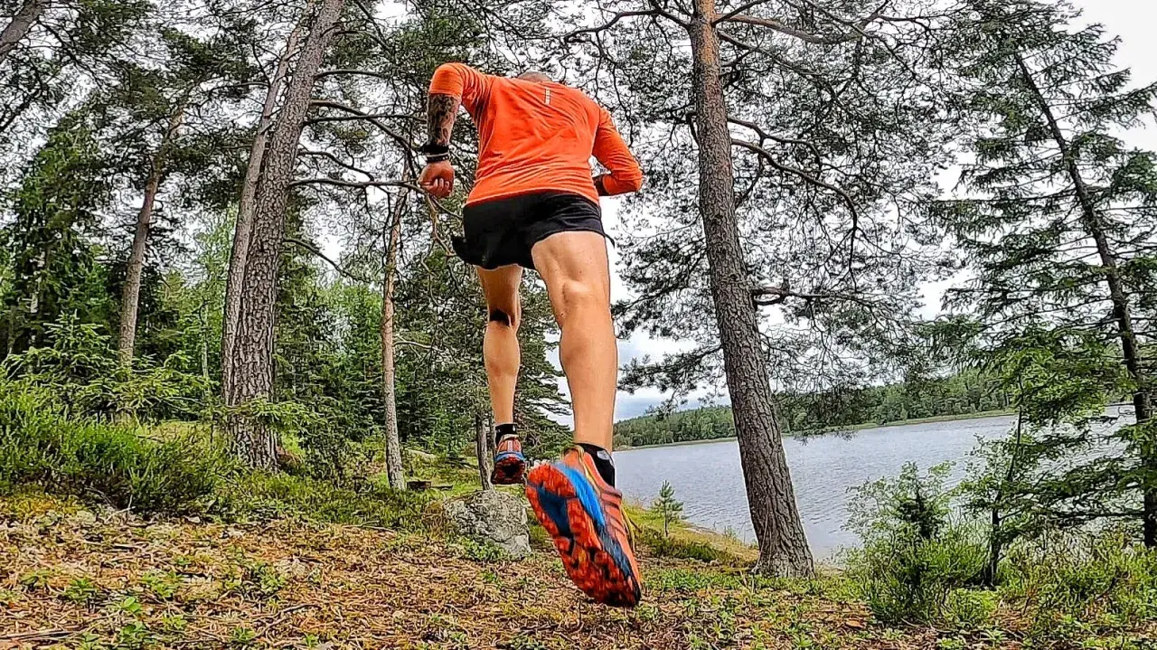 En löpare i orange tröja och svarta shorts springer längs en sjö i skogen. En del av the run Uppsala.