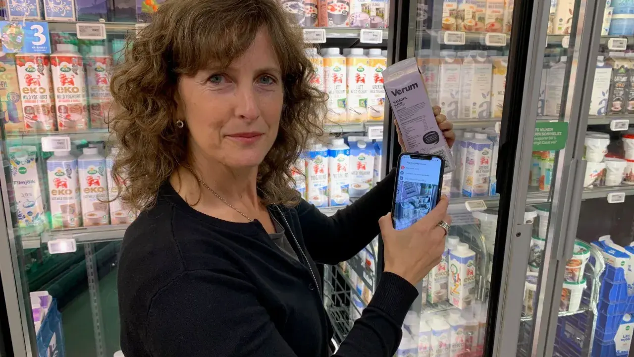 Kvinna håller upp en mjölkförpackning och en mobiltelefon, visar hur man kollar vad som innehåller socker. Kylskåp med mejeriprodukter i bakgrunden.
