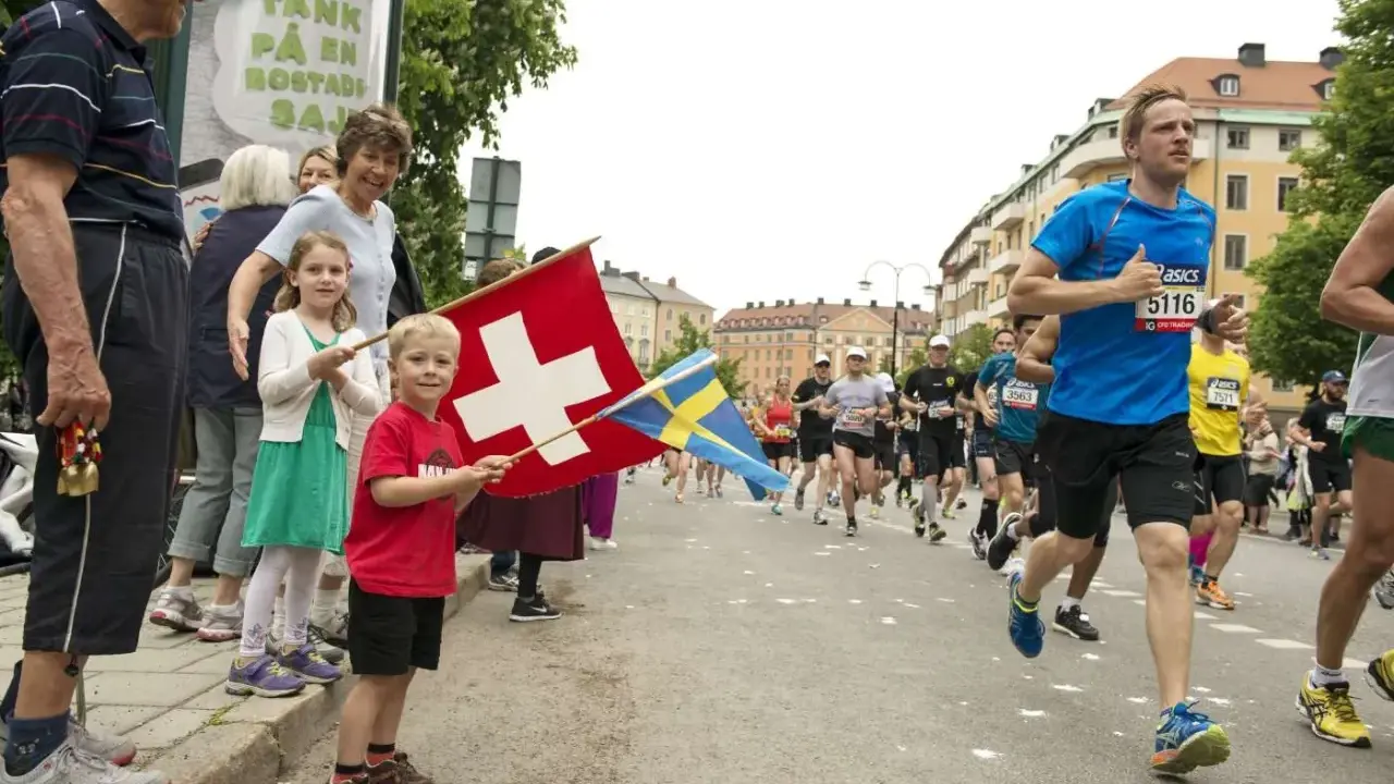 Barn viftar med schweizisk och svensk flagga vid ett lopp. Löpare på marathon distans passerar.