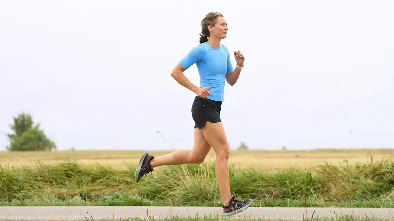 Kvinna i blå tröja och svarta shorts springer på en landsväg. Hon har en imponerande **run streak** och njuter av löprundan i det öppna landskapet.
