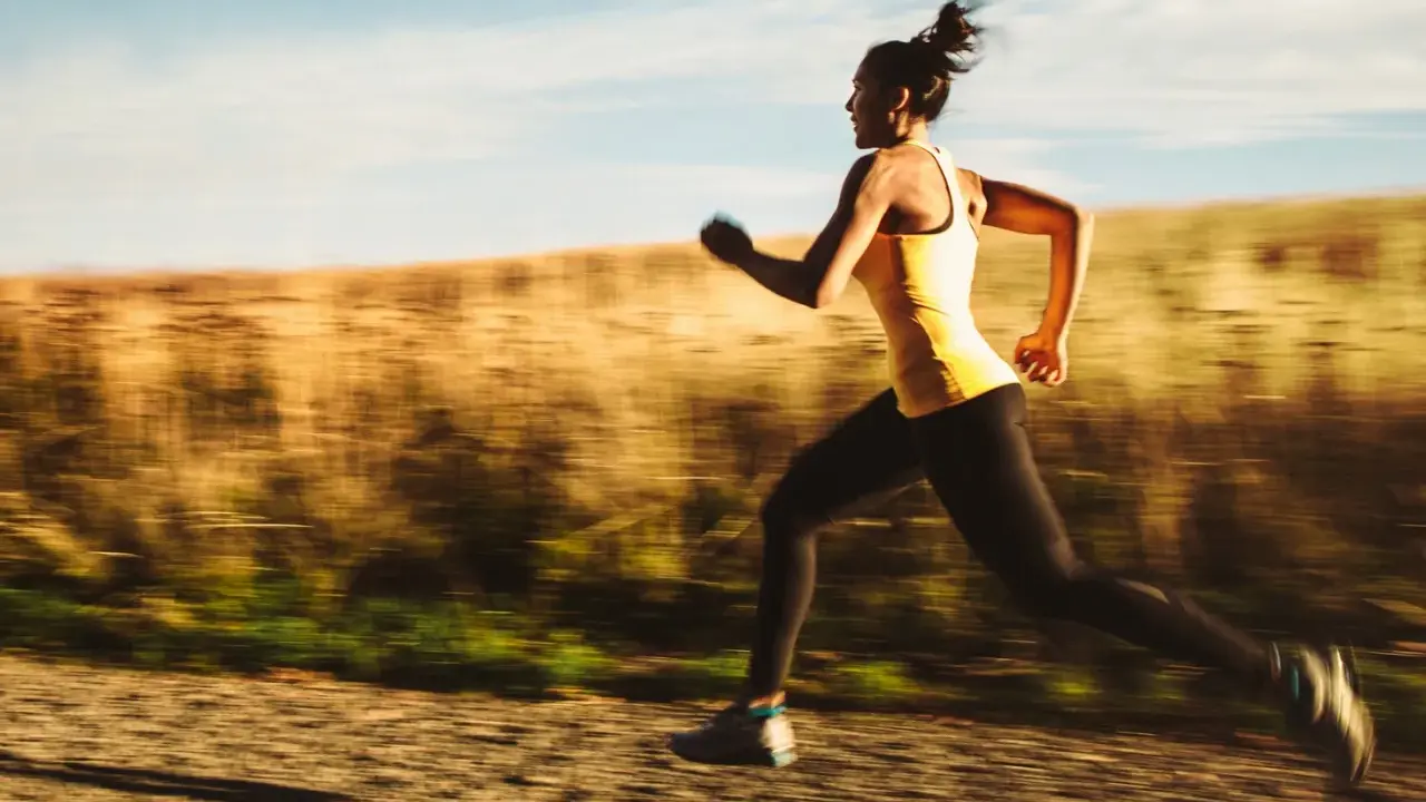 Kvinna i gult linne och svarta tights springer snabbt på en grusväg, en del av ett intensivt hiit pass.