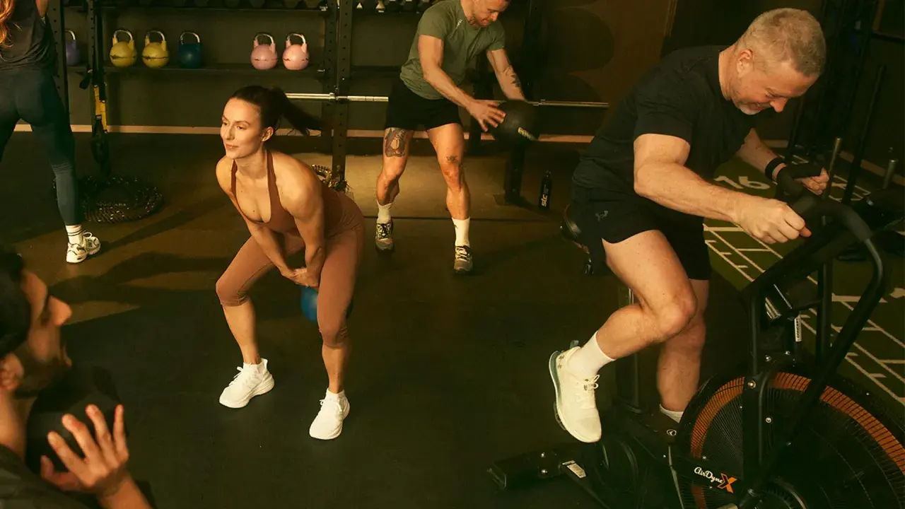 Träningspass med fokus på kondition och styrka. En kvinna gör knäböj med kettlebell, en man kastar medicinboll och en annan cyklar på en Air Bike. Kanske en övning för att hålla gunde svan vilopuls nere.