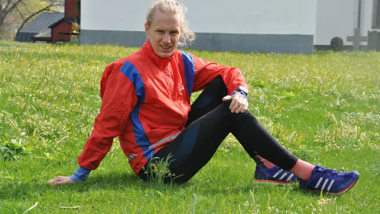 En löpare i röd jacka och svarta tights vilar i gräset, redo att springa milen under 50 minuter.