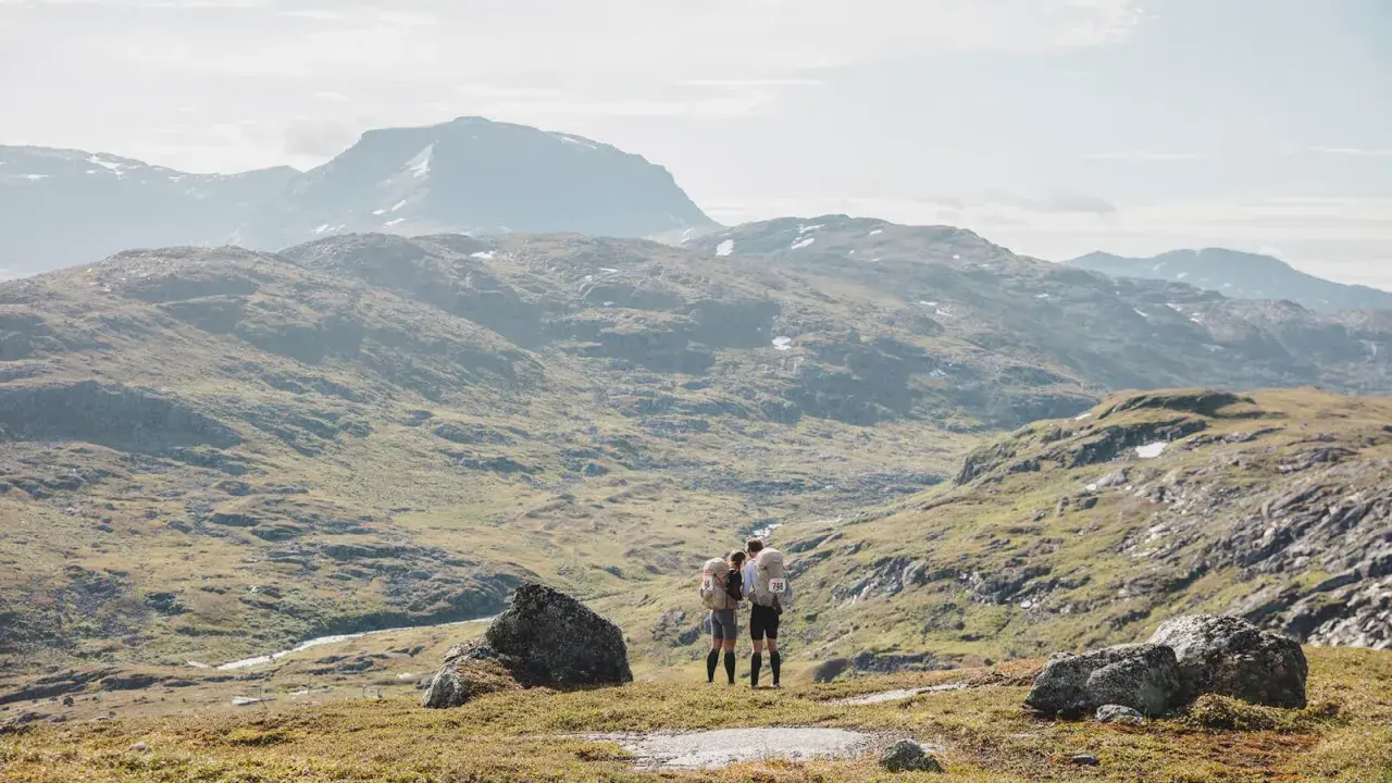 Två vandrare med bamm utrustning står på en bergssluttning med vidsträckt utsikt över fjällandskap.
