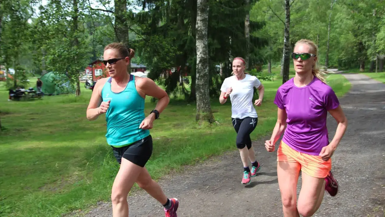 Tre kvinnor springer på en grusväg i en skogsmiljö, zon 2.