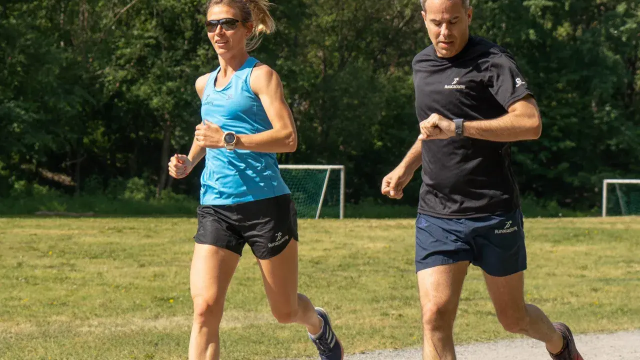 Two runners, a woman in blue and a man in black, run on a gravel path with a grassy field and trees in the background.