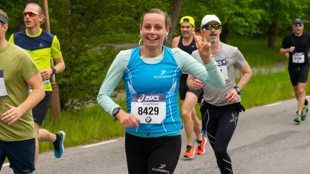 Glada löpare i ett 10 km lopp. En kvinna med nummer 8429 ler och gör segertecken.