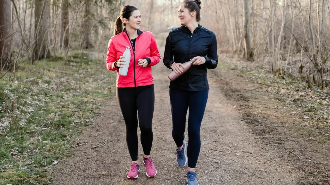 Two women in athletic wear walk on a forest path, holding water bottles.