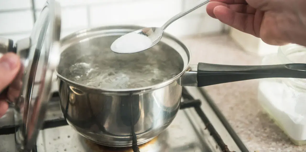 En sked med salt tillsätts i kokande vatten. En del av locket är upplyft. En burk med salt syns i bakgrunden, redo för egen resorb.