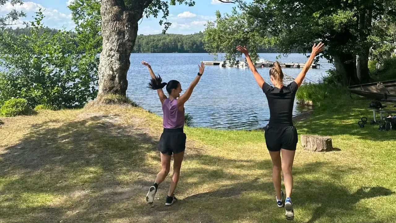 Två kvinnor hoppar glatt vid en sjö, redo att springa en mil om dagen i den vackra svenska naturen.