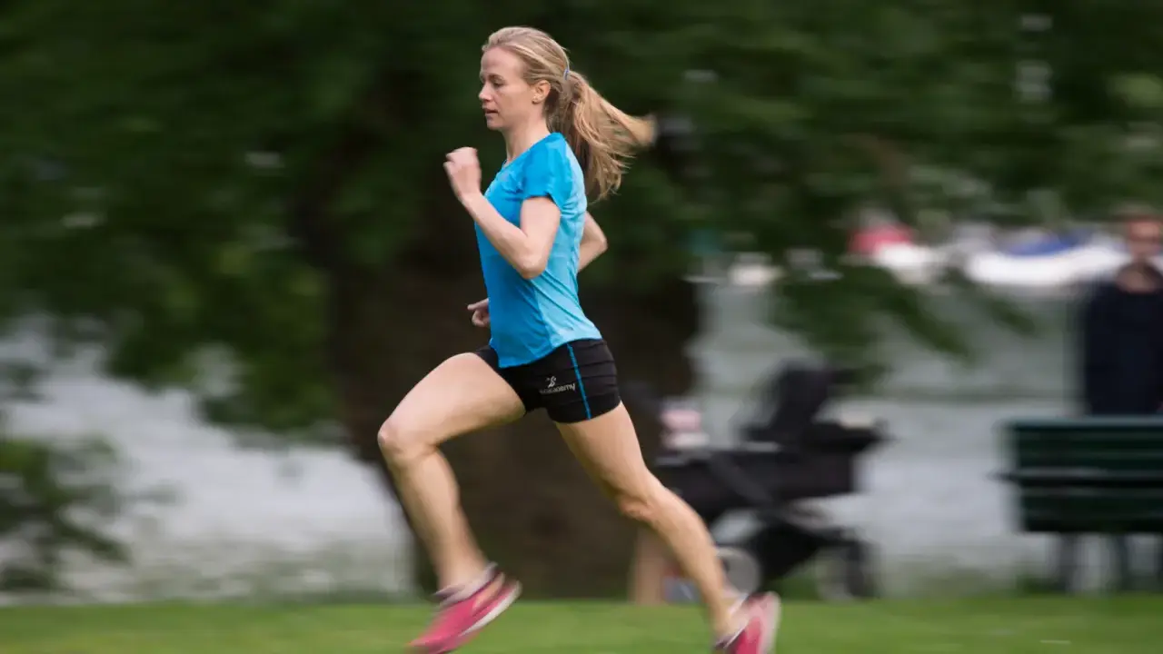 En person som springer snabbt över en grön gräsmatta, med träd och en sjö i bakgrunden.