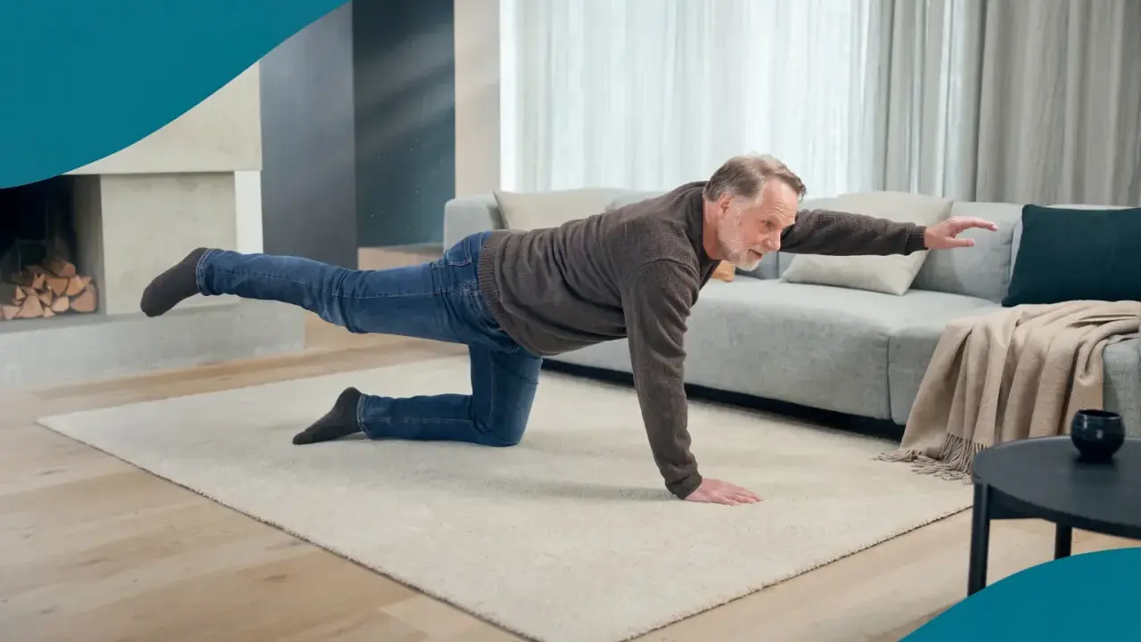 Äldre man tränar balans på en matta i vardagsrummet. Han sträcker ut ett ben och en arm, och får verkligen **hålla i magen** för att inte tappa balansen.