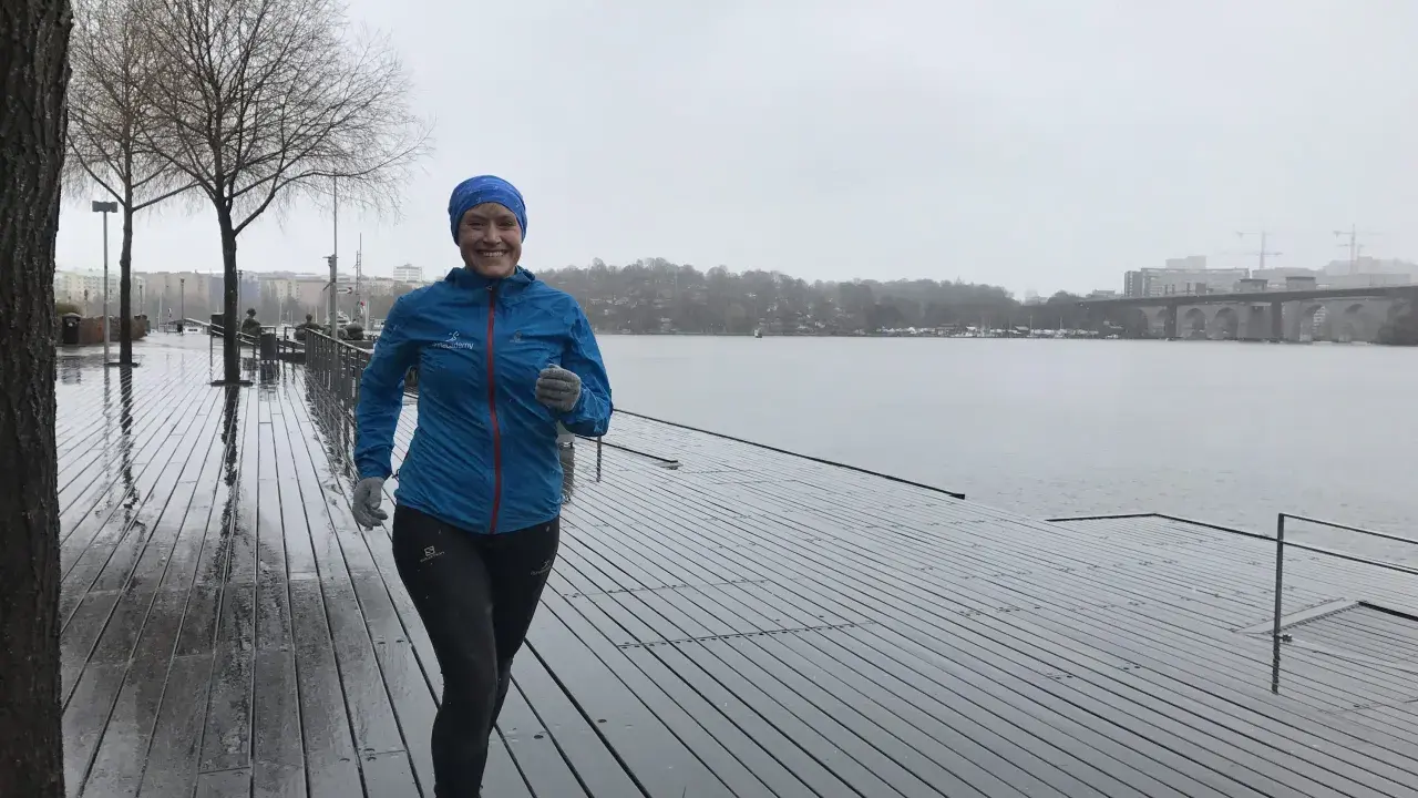 Glad kvinna med blå jacka och mössa springer i regn längs en träbrygga vid en sjö.