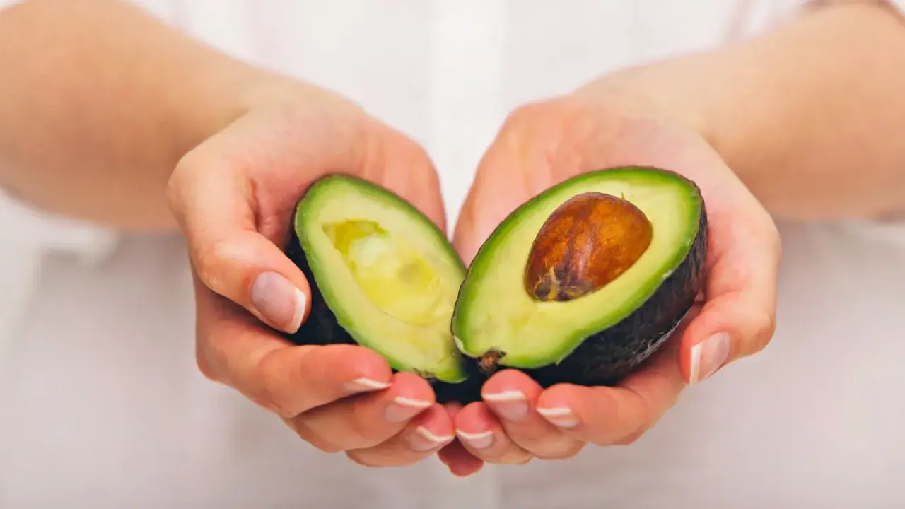 Händerna håller en avokado, delad på mitten, som visar dess gröna kött och bruna kärna. En näringsrik källa till avokado vitaminer.