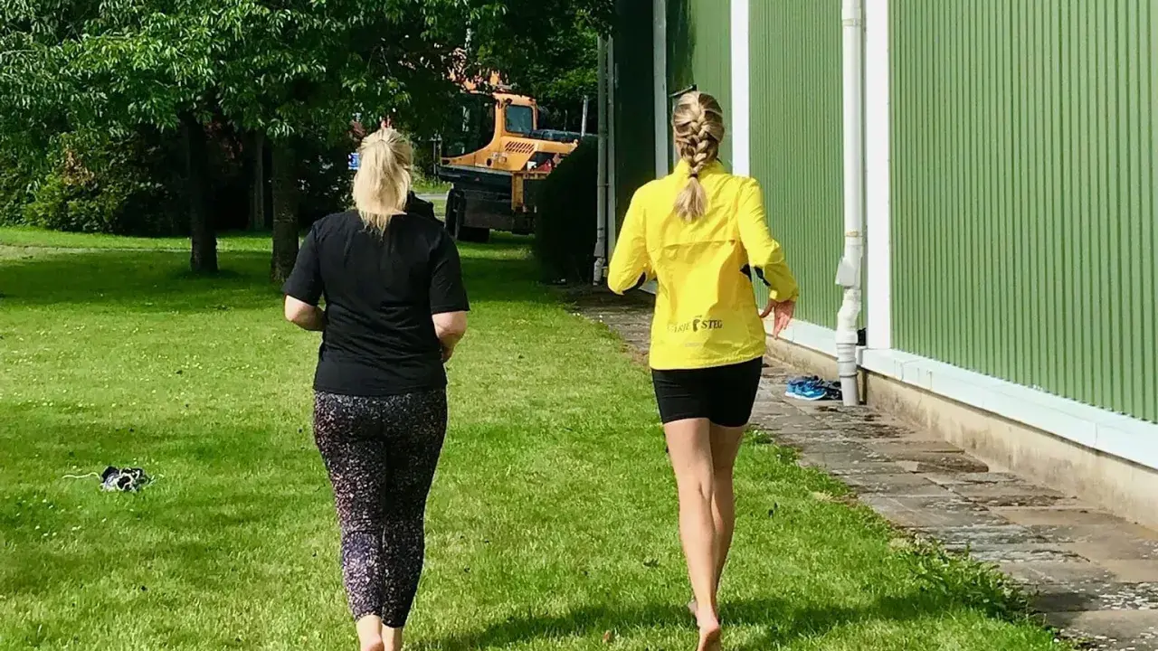 Två kvinnor njuter av en löprunda barfota på gräset en solig dag.