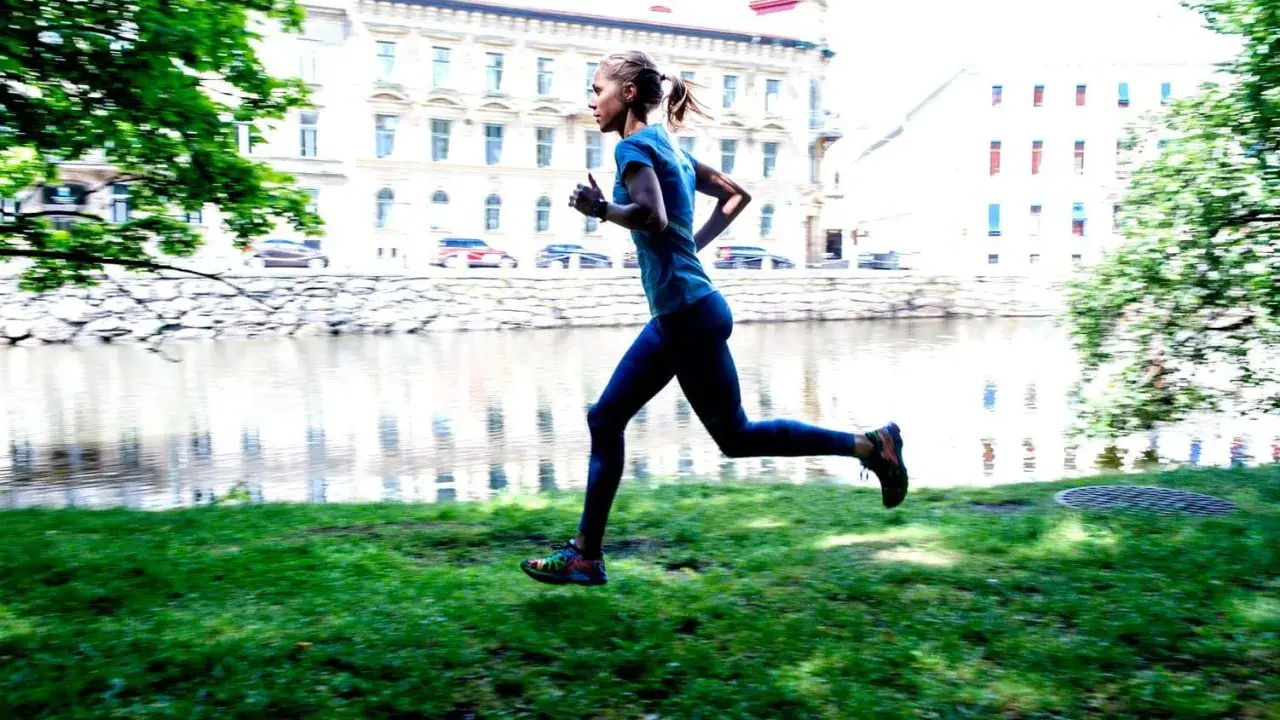 En kvinna joggar långsamt eller går snabbt längs en gräsmatta vid en flod, med byggnader i bakgrunden.