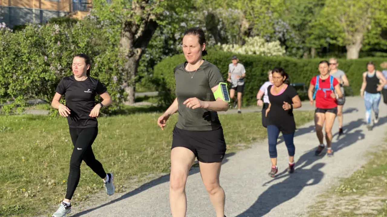En grupp löpare på en grusväg, kanske på väg att klara 1 mil. Solen skiner och träden är gröna.