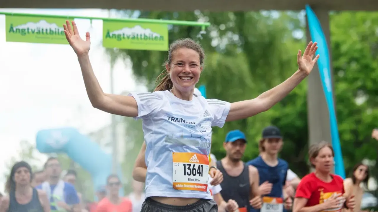 Glad kvinna med armarna uppe vid målgången i ett maraton. En lycklig löpare som njuter av sin maraton tid.