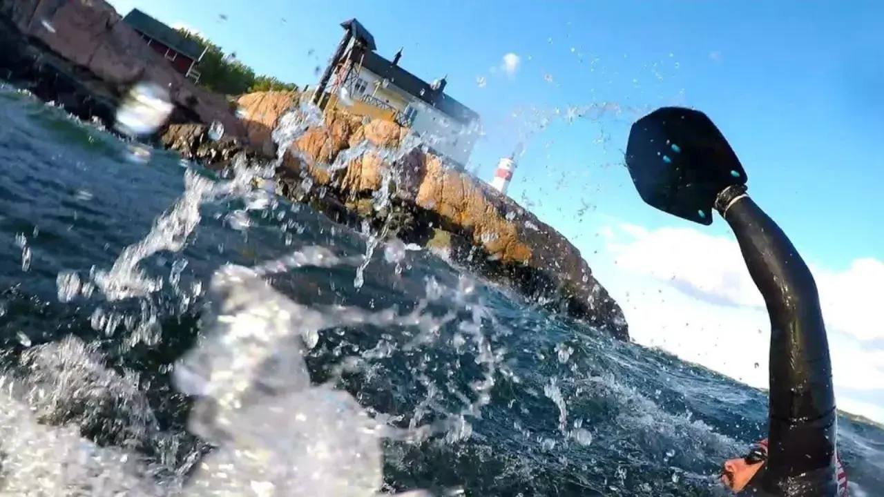 Vattenstänk och en paddel i förgrunden. I bakgrunden en klippa med hus och ett litet fyrhus. En del av en farleden swimrun-deltagare syns.
