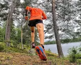 En löpare i orange tröja och svarta shorts springer längs en sjö i skogen. En del av the run Uppsala.