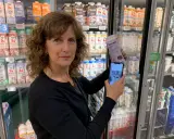 Kvinna håller upp en mjölkförpackning och en mobiltelefon, visar hur man kollar vad som innehåller socker. Kylskåp med mejeriprodukter i bakgrunden.