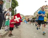 Barn viftar med schweizisk och svensk flagga vid ett lopp. Löpare på marathon distans passerar.
