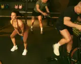 Träningspass med fokus på kondition och styrka. En kvinna gör knäböj med kettlebell, en man kastar medicinboll och en annan cyklar på en Air Bike. Kanske en övning för att hålla gunde svan vilopuls nere.