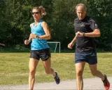Two runners, a woman in blue and a man in black, run on a gravel path with a grassy field and trees in the background.
