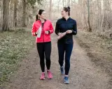 Two women in athletic wear walk on a forest path, holding water bottles.