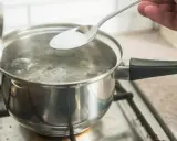 En sked med salt tillsätts i kokande vatten. En del av locket är upplyft. En burk med salt syns i bakgrunden, redo för egen resorb.