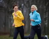 Två kvinnor springer i parken, förberedda för ett halv maraton. Den ena bär en gul jacka, den andra en blå.