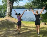 Två kvinnor hoppar glatt vid en sjö, redo att springa en mil om dagen i den vackra svenska naturen.