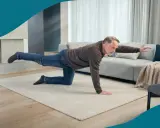 Äldre man tränar balans på en matta i vardagsrummet. Han sträcker ut ett ben och en arm, och får verkligen **hålla i magen** för att inte tappa balansen.