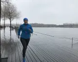 Glad kvinna med blå jacka och mössa springer i regn längs en träbrygga vid en sjö.
