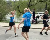 Två löpare ger varandra high-five under roliga lopp. En kvinna i blått och en man i blått. I bakgrunden syns en barnvagn och en sjö.