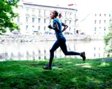 En kvinna joggar långsamt eller går snabbt längs en gräsmatta vid en flod, med byggnader i bakgrunden.
