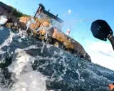 Vattenstänk och en paddel i förgrunden. I bakgrunden en klippa med hus och ett litet fyrhus. En del av en farleden swimrun-deltagare syns.