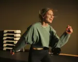 Glad kvinna med blont hår som springer intervaller i ett gym. Hon har på sig en grön tröja och svarta träningsbyxor.