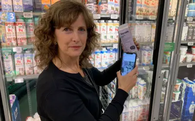 Kvinna håller upp en mjölkförpackning och en mobiltelefon, visar hur man kollar vad som innehåller socker. Kylskåp med mejeriprodukter i bakgrunden.