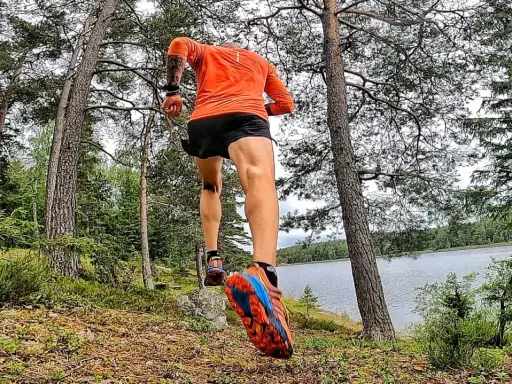 En löpare i orange tröja och svarta shorts springer längs en sjö i skogen. En del av the run Uppsala.