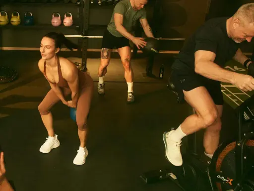 Träningspass med fokus på kondition och styrka. En kvinna gör knäböj med kettlebell, en man kastar medicinboll och en annan cyklar på en Air Bike. Kanske en övning för att hålla gunde svan vilopuls nere.
