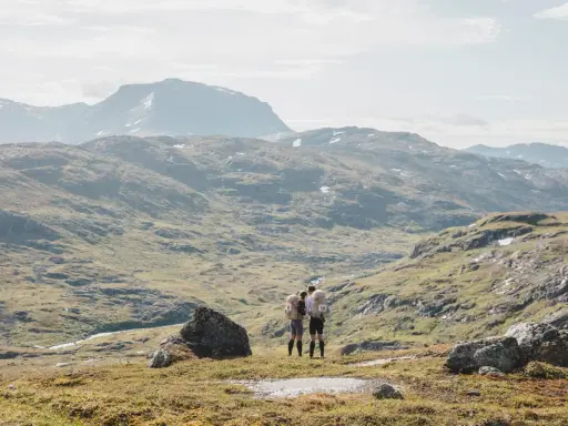 Två vandrare med bamm utrustning står på en bergssluttning med vidsträckt utsikt över fjällandskap.
