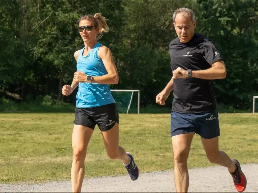 Two runners, a woman in blue and a man in black, run on a gravel path with a grassy field and trees in the background.