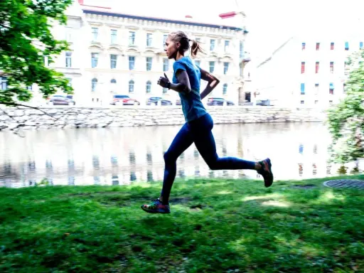 En kvinna joggar långsamt eller går snabbt längs en gräsmatta vid en flod, med byggnader i bakgrunden.