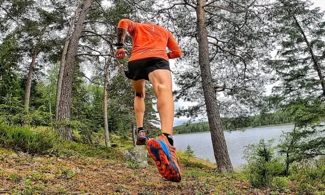 En löpare i orange tröja och svarta shorts springer längs en sjö i skogen. En del av the run Uppsala.