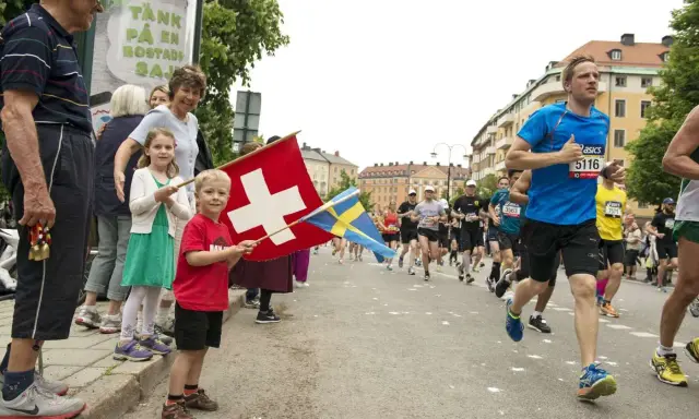 Barn viftar med schweizisk och svensk flagga vid ett lopp. Löpare på marathon distans passerar.