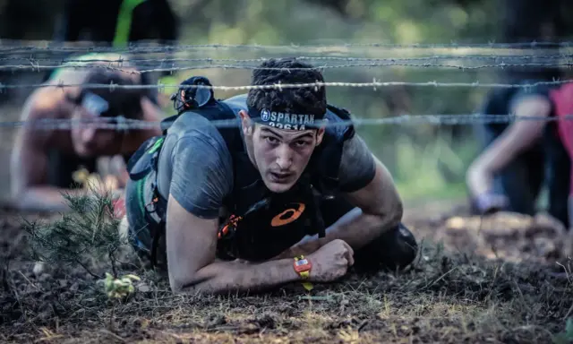 En deltagare i ett ocr sport-lopp kryper under taggtråd.