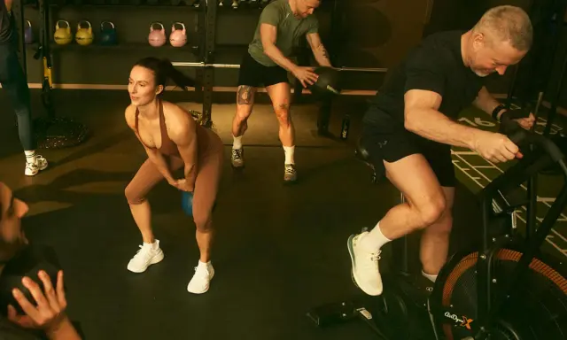 Träningspass med fokus på kondition och styrka. En kvinna gör knäböj med kettlebell, en man kastar medicinboll och en annan cyklar på en Air Bike. Kanske en övning för att hålla gunde svan vilopuls nere.