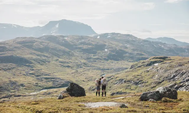 Två vandrare med bamm utrustning står på en bergssluttning med vidsträckt utsikt över fjällandskap.