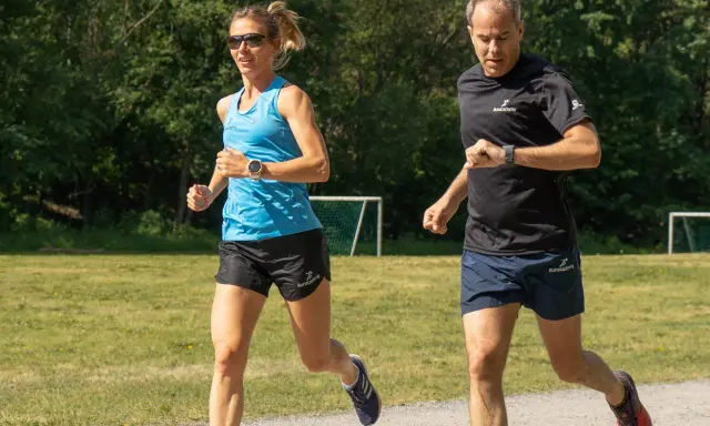 Two runners, a woman in blue and a man in black, run on a gravel path with a grassy field and trees in the background.